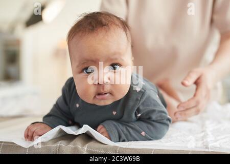 Nahaufnahme Porträt von niedlichen gemischt Rennen Baby Blick auf die Kamera, während auf Wickeltisch mit verschwommener Form der fürsorglichen Mutter im Hintergrund liegend, Kopie SPAC Stockfoto