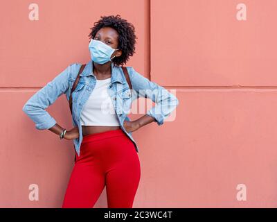 Positive afro hübsches Mädchen, lächeln mit den Augen und tragen schützende und medizinische Maske, weißes T-Shirt und Jeans Jacke, rote Hose über Pastell Koralle Pin Stockfoto