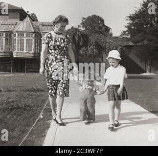 1950er Jahre, historisch, "erste Schritte", draußen auf einem Weg in das Gelände eines alten Hauses, eine Mutter, die die ihres Kleinkindes hält, während ihre junge Tochter die andere Hand des Kleinkindes hält, während sie ihre ersten Schritte macht, stehend, England, Großbritannien. Stockfoto