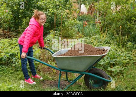 Ein Mädchen in einer leuchtend rosa Jacke versucht, eine schwere Schubkarre mit Sand zu bewegen. Stockfoto