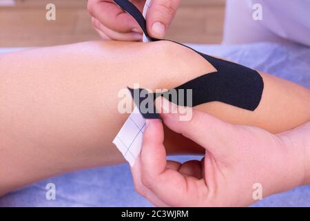 Hände einer nicht erkennbaren Physiotherapeutin, die schwarzes Klebeband auf das Knie einer Frau legt Stockfoto