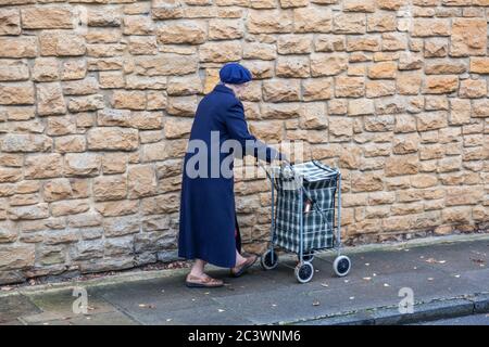 Eine ältere Dame mit Mantel und Baskenmütze schiebt ihren Spaziergänger oder Einkaufskorbus an einer Kalksteinwand vorbei, Dorset, England. Stockfoto