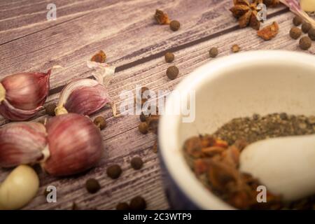 Piment in einem Keramikmörtel und verstreute Knoblauchzehen, Sternanis und Piment auf einem Holztisch. Nahaufnahme Stockfoto