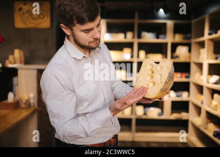 Hansome Mann hält große Scheibe Käse maasdam in der Hand. Käse mit großen Löchern. Hintergrund der Regale mit Käse Stockfoto