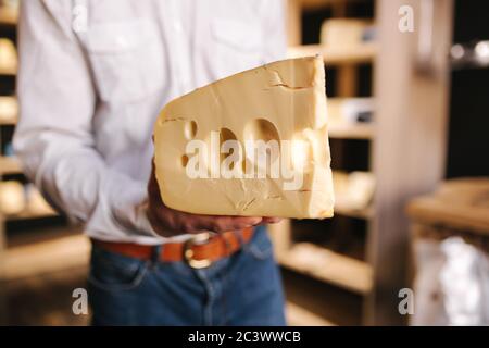 Hansome Mann hält große Scheibe Käse maasdam in der Hand. Käse mit großen Löchern. Hintergrund der Regale mit Käse Stockfoto