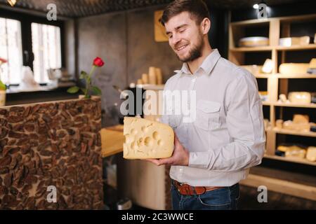 Hansome Mann hält große Scheibe Käse maasdam in der Hand. Käse mit großen Löchern. Hintergrund der Regale mit Käse Stockfoto