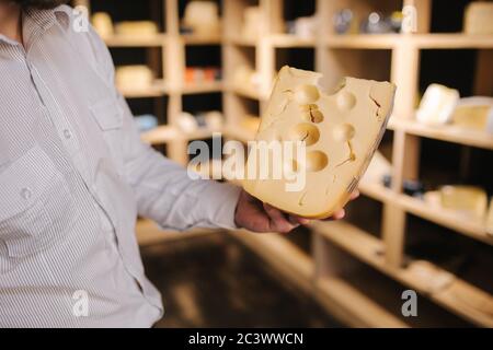 Hansome Mann hält große Scheibe Käse maasdam in der Hand. Käse mit großen Löchern. Hintergrund der Regale mit Käse Stockfoto