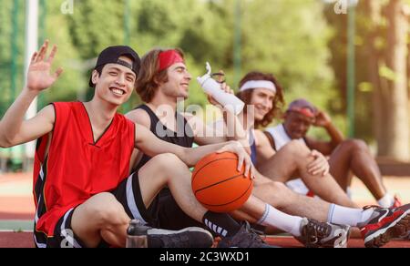 Asiatischer Spieler und sein multinationales Team, die sich auf der Basketballarena im Freien ausruhen, leeren Raum Stockfoto