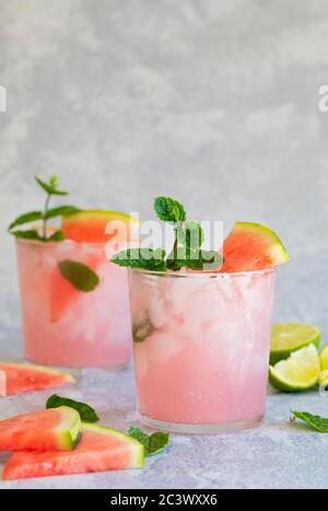 Zwei rosa Getränke mit Wassermelone und Limette, mit Minzgarnierung. Das Getränk ist auf einem grauen Hintergrund, der mit zerdrücktem Eis verstreut ist. Vertikales Foto mit Stockfoto