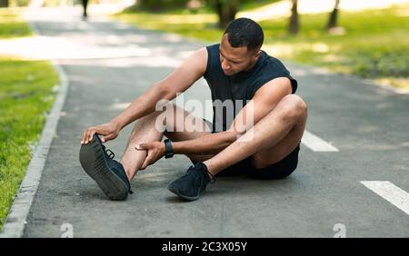 Sportverletzung. Afroamerikanischer Läufer sitzt auf der Joggingstrecke und fühlt Schmerzen in seinem Knöchel Stockfoto