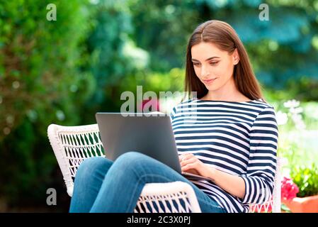 Aufnahme einer schönen jungen Frau mit ihrem Laptop, während sie im Garten sitzt und zu Hause arbeitet. Stockfoto