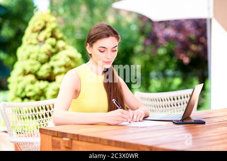 Porträtaufnahme einer schönen Frau, die etwas schreibt, während sie am Schreibtisch im Garten sitzt. Stockfoto