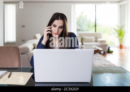 Geschäftsfrau, die mit dem Smartphone spricht und zuhause einen Laptop benutzt. Remote Working Konzept. Stockfoto
