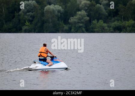 Man in Life Weste reitet einen Wasserscooter auf dem Fluss auf Waldgrund. Sicherheit auf dem Wasser während der Sommerferien Stockfoto