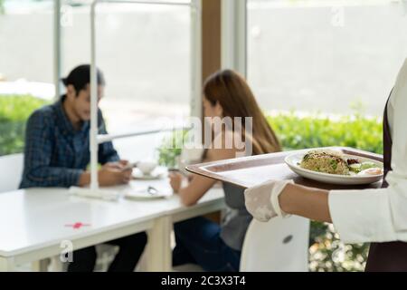 Rückansicht Asian Kellnerin hält Food Tablett zu servieren Mahlzeit an Kunden mit Kunden im Hintergrund. Neues normales Restaurant Lifestyle Konzept. Stockfoto
