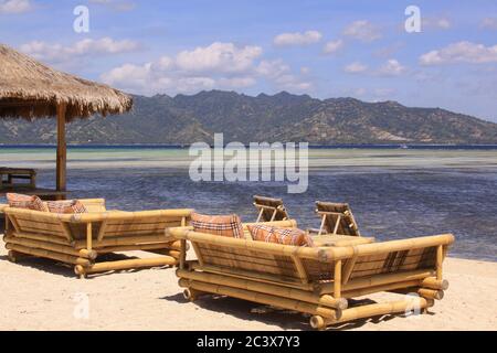 Tropischer Strohschirm und Sofas mit Kissen an der Strandbar am sonnigen exotischen Strand mit Blick auf die Berge auf Gili Air Island, Lombok, Indonesien Stockfoto