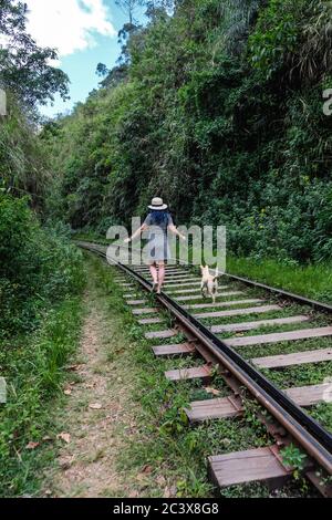 Ein Mädchen im Hut, das mit einem Hund entlang der Eisenbahnstrecke läuft. Grüner Wald draußen. Warten auf den Ella Zug. Stockfoto