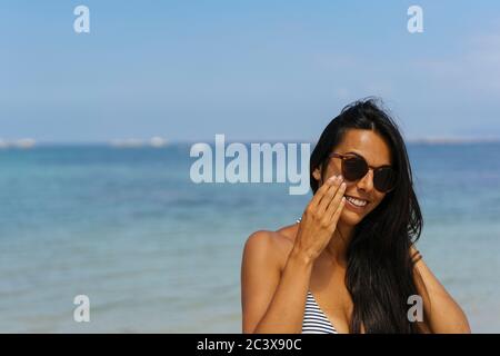 Lächelnde Frau, die Sonnenschutzcreme auf ihr Gesicht am Strand aufgibt Stockfoto