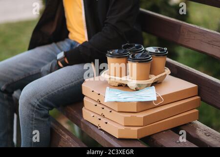 Courier in Schutzmaske und medizinischen Handschuhen liefert Pizza, Kaffee und Schutzmaske. Delivery Boy sitzen auf Bank und warten auf Kunden. Lieferung Stockfoto