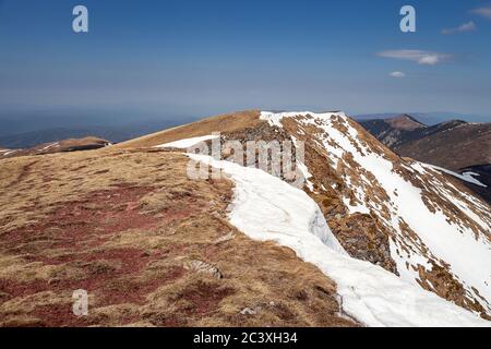 Schöne Aussicht von einem Berg Hügel bedeckt von verbleibendem Schnee Am Ende des Winters und entfernten Berggipfeln unter Ein blauer Himmel Stockfoto