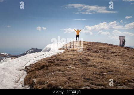 Junger Bergwanderer mit gelber Jacke auf dem Gipfel Feiern mit seinen Händen in der Luft und schauen Die Entfernung Stockfoto