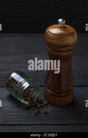 Glas mit schwarzem Pfeffer und einer Mühle auf schwarzem Holzgrund Stockfoto