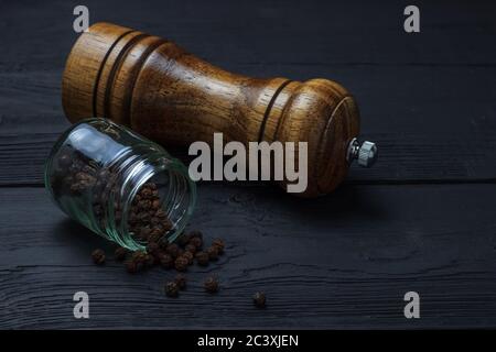 Glas mit schwarzem Pfeffer und einer Mühle auf schwarzem Holzgrund Stockfoto