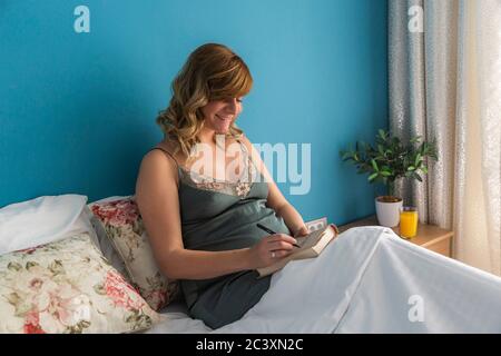 Eine schwangere lächelnde Frau in einer halbsitzenden Position in ihrem Bett, während sie etwas auf einem Notizbuch schreibt. Stockfoto