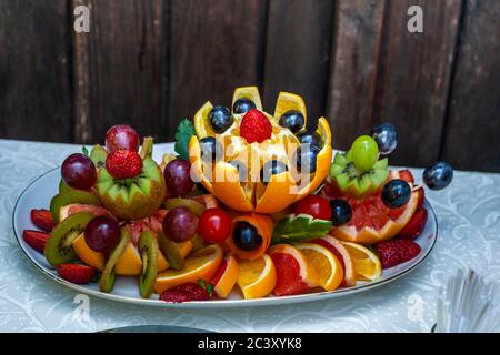 Orangen, die zu Blumenformen geschnitzt sind, zusammen mit Kiwi, Trauben und Erdbeeren, bilden eine aufwendige Obstarrangement auf dem Buffet. Schwarze Trauben und eine Erdbeerkrone bekrönen die zentrale Blume, während Grapefruit-Keile den Rand des Tellers säumen. Aufgenommen bei einer bulgarischen Hochzeit in Dobrich, Bulgarien. Stockfoto