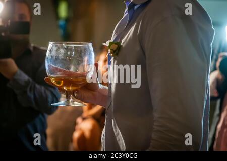Ein Gast mit einer weißen Blume an seinem Hemd hält ein bauchiges Cognacglas mit bernsteinfarbener Flüssigkeit. Ein Fotograf fängt die Feier im Hintergrund ein. Aufgenommen an einer bulgarischen Strandhochzeit in Albena, Gemeinde Balchik, Bulgarien. Stockfoto