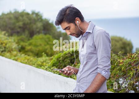 Empfangen einer Textnachricht. Handsome man in Grey Shirt Halten und Blick auf Handy im Freien, grünen Busch, Meer auf Hintergrund. Stockfoto