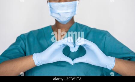 Afrikanische Krankenschwester tragen Gesichtsmaske, Handschuhe, blaue Uniform zeigt Herz Hände Form. Stockfoto