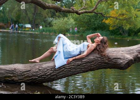 Ein schönes Mädchen mit lockigen Haaren in einem blauen Kleid ist Auf einem großen Baum liegend vor dem Hintergrund eines Park mit einem See Stockfoto
