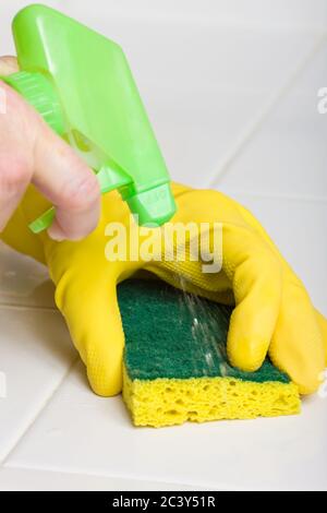 Frau trägt einen gelben Gummihandschuh und wäscht eine Küchenarbeitsplatte mit einem Schwamm und einem Spray, das aus einer Reinigungsflasche kommt Stockfoto