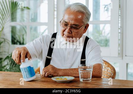 Senior Asian Mann mit Tasse Wasser und Pille Einnahme Heilmittel in gemütlichen Raum zu Hause. Alter Mann nimmt Medizin Stockfoto