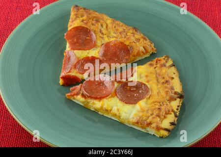 Zwei Scheiben gebackene Pepperoni Pizza auf blauem Teller auf roter Tischdecke Stockfoto