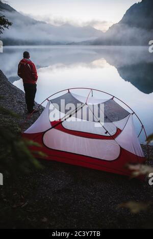 Österreich, Plansee, junger Mann, der in der Nähe des Zeltes steht und den Plansee bei Sonnenaufgang betrachtet Stockfoto