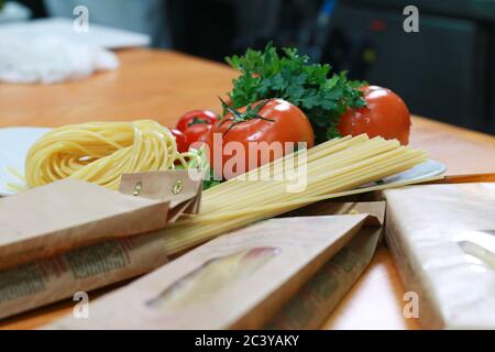 Ungekochte Nudeln in einer Papierverpackung mit frischen Tomaten und Sellerie. Frische Spaghetti-Zutaten auf dem Holztisch. Stockfoto