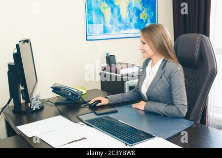 Die Dame, die in einem modernen Stil in einem formellen Anzug am Schreibtisch aufsteht, schaut auf den Computermonitor. Stockfoto