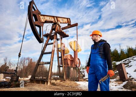 Horizontale Momentaufnahme eines Ölarbeiters in orangefarbenem Helm während seines Arbeitstages in einem Ölfeld, hält ein Ölingenieur einen Rohrschlüssel und schaut auf einen Pumpenheber hinter ihm. Konzept der Öl- und Gasindustrie Stockfoto