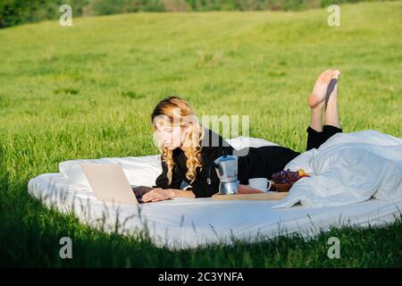 Frau, die gerne am Laptop arbeitet, im Bett im Freien liegt, unter sanfter Frühlingssonne Stockfoto