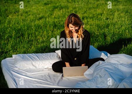 Zufriedene, zufriedene Frau, die auf einem Laptop arbeitet, im Bett im Freien sitzend Stockfoto