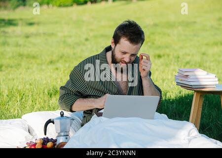 Mann, der im Bett im Freien in grassfield sitzt und an seinem Laptop arbeitet Stockfoto