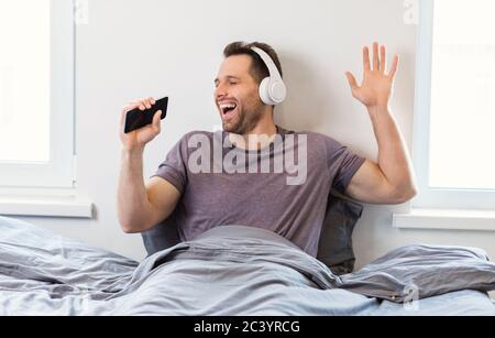 Mann Trägt Kopfhörer Singing Song Sitzen Im Bett Zu Hause Stockfoto