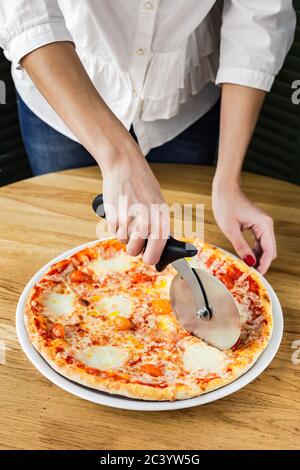 Schneiden Pizza margherita mit einem Pizzaschneider, serviert auf einem runden Holzbrett, natürliches Licht Stockfoto
