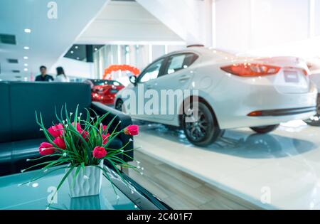 Fake rote Blumen in Keramik weiße Vase auf verschwommenem Hintergrund des Verkaufs Person beschäftigt sich mit Kunden in modernen Showroom. Autohaus. Verschwommenes Luxusauto Stockfoto