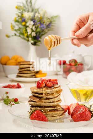 Pfannkuchen mit Erdbeeren, leckeres Frühstück. Eine Frau gießt Honig auf einen Stapel Pfannkuchen. Instagram vertikale Lebensmittel Foto Stockfoto