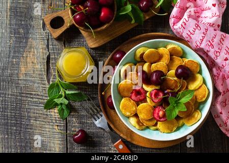 Winziges Pfannkuchen-Frühstück. Mini-Pfannkuchen mit Kirschen und Honig auf rustikalem Holztisch. Draufsicht flach lagen Hintergrund. Speicherplatz kopieren. Stockfoto