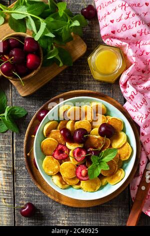 Winziges Pfannkuchen-Frühstück. Mini-Pfannkuchen mit Kirschen und Honig auf rustikalem Holztisch. Draufsicht flach lagen Hintergrund. Speicherplatz kopieren. Stockfoto