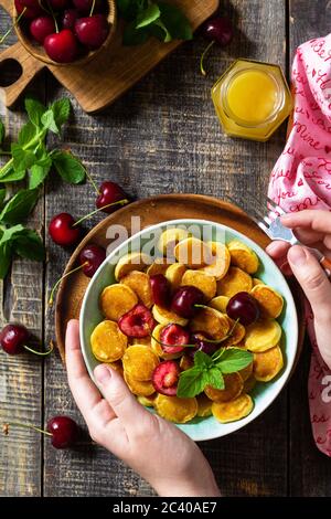 In den Händen einer Frau Frühstück. Mini-Pfannkuchen mit Kirschen und Honig auf einem rustikalen Holztisch. Draufsicht flach lagen Hintergrund. Stockfoto